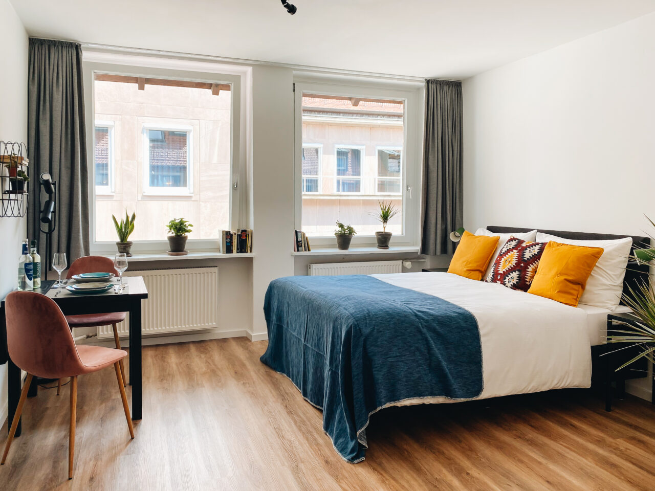 Bett mit blauer Tagesdecke und senfgelben Kissen vor zwei Fenstern