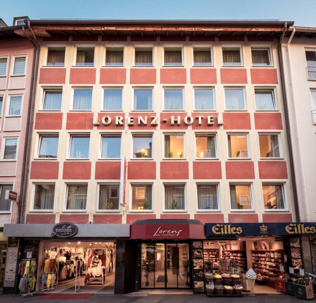 Fassade des Hauses mit roten Flächen, dem Eingang der Lorenz Apartments und Geschäften im Erdgeschoss